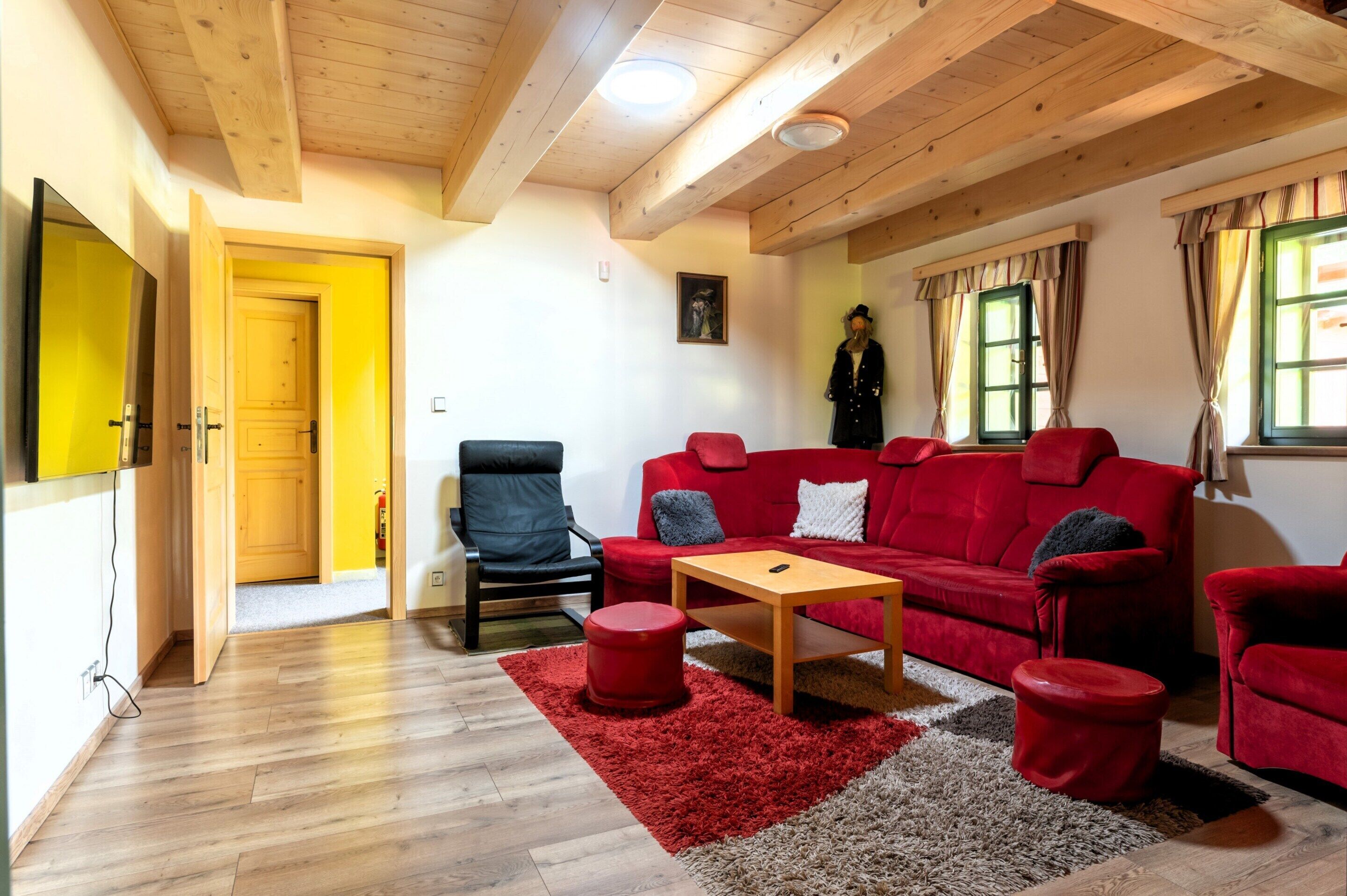 A cozy living room with a red sofa, a wooden coffee table, and a red ottoman. A black chair is positioned near the sofa, and a wooden door leads to another room.