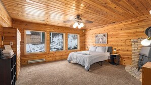 Ein gemütliches Schlafzimmer in einer Holzhütte mit Bett, Nachttisch, Heizung und großen Fenstern mit Blick auf winterliche Landschaften.