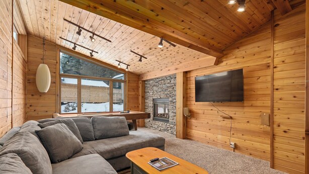 Ein gemütliches Blockhaus-Interieur mit einem Ecksofa, einem Holztisch, einem Kamin aus Stein und einem großen Fenster mit Blick auf die verschneite Landschaft.