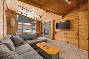Een gezellig interieur van een vakantiehuis van hout met een hoekbank, een houten salontafel, een stenen open haard en een groot raam met uitzicht op het besneeuwde landschap.