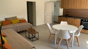 A modern living room with a sofa, a small table, and a dining area with chairs. There is a kitchen with wooden cabinets and a stove.