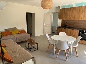 A modern living room with a sofa, a small table, and a dining area with chairs. There is a kitchen with wooden cabinets and a stove.