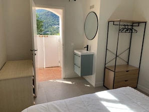 A bedroom with a bed, a bedside table, a wardrobe, a mirror, a sink, and a view of a mountainous landscape through an open door.