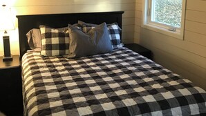 Ein Schlafzimmer mit kariertem Bettbezug, einem Nachttisch mit Lampe und einem Fenster mit Blick auf den Garten.