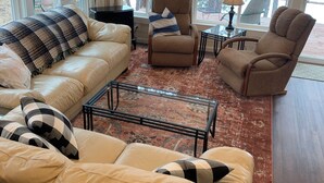 Ein gemütliches Wohnzimmer mit einer Couch, Sesseln, einem Couchtisch und einem Fernseher. Große Fenster bieten Blick auf Bäume.