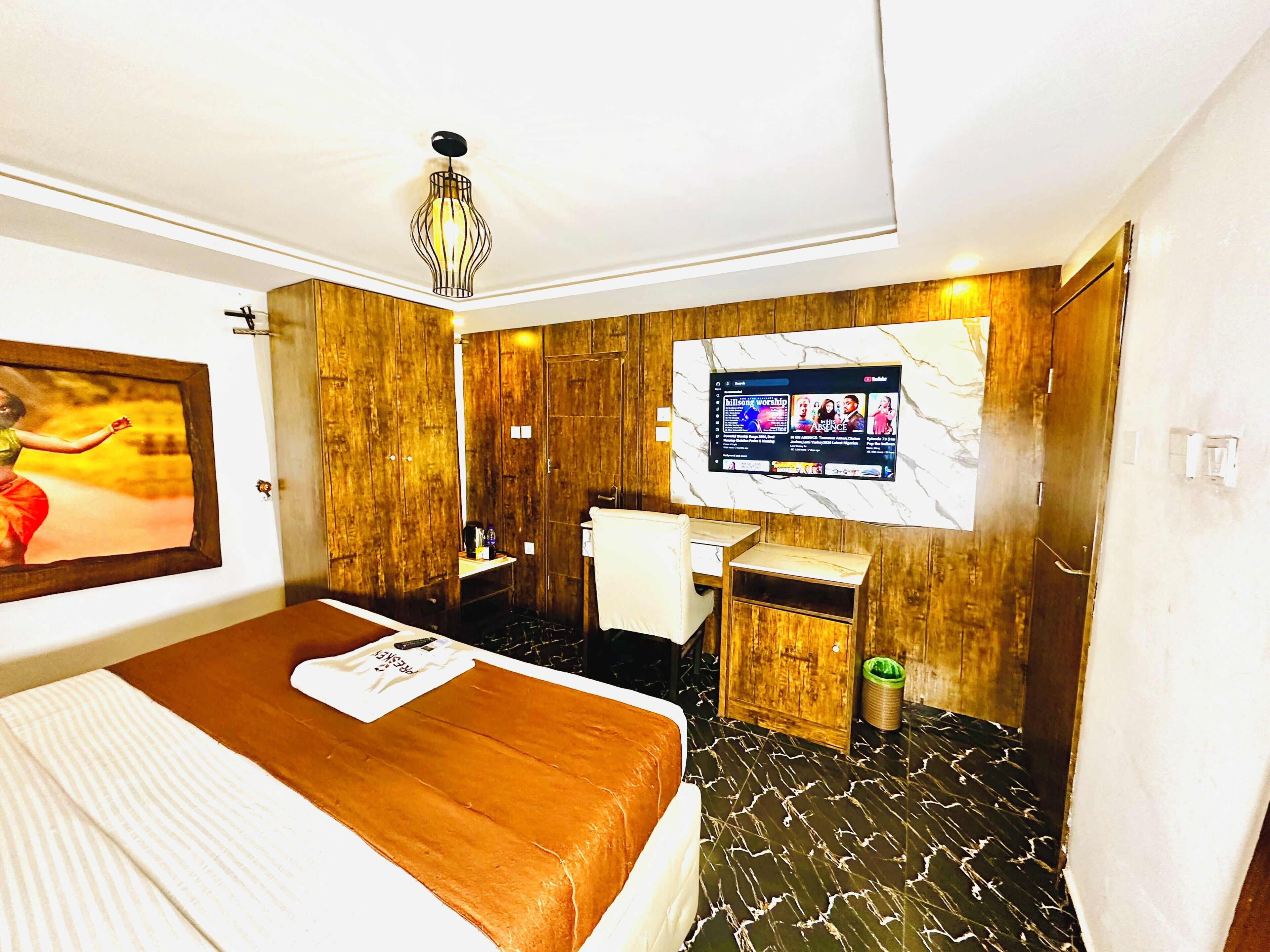 A hotel room with a bed, a television mounted on the wall, a wooden wardrobe, and a small bathroom area.