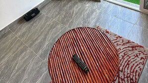 Une pièce avec un téléviseur fixé au mur, une plante, une télécommande posée sur une table ronde, et une vue sur un balcon aménagé avec du mobilier d’extérieur.