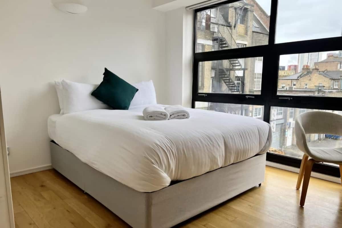 A modern hotel room with a large bed, white bedding, a green pillow, and a view of the city through a large window.