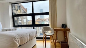 Hotellihuone, jossa on suuri ikkuna, sänky, tuoli, työpöytä ja patteri.