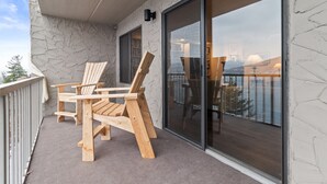 A balcony with a wooden table and chairs, a view of the cityscape through large glass doors, and a stone wall.