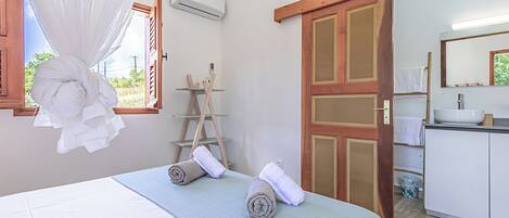 A bedroom with a bed, a window with sheer curtains, a wooden ladder, and a bathroom visible through an open door.