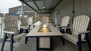 An outdoor patio with a long table and six Adirondack chairs, a fire pit in the center, and a covered area with a metal roof. - Fish Stories 5 Bedroom/3.5 Bathroom With Pool and Playscape (Port O'Connor)