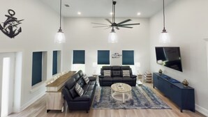 A modern living room with a sofa, coffee table, and TV. There are ceiling fans, pendant lights, and a large window with blinds. - Fish Stories 5 Bedroom/3.5 Bathroom With Pool and Playscape (Port O'Connor)