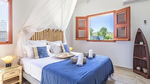 A neatly arranged bedroom with a bed, a bedside table, a nightstand, a window with open shutters, and a bookshelf.