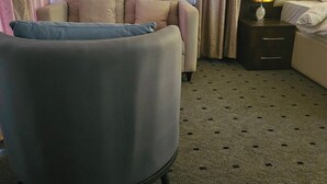 A hotel room with a sofa, a chair, a bed, a nightstand, and a lamp. - Anibex Luxe (Abuja)