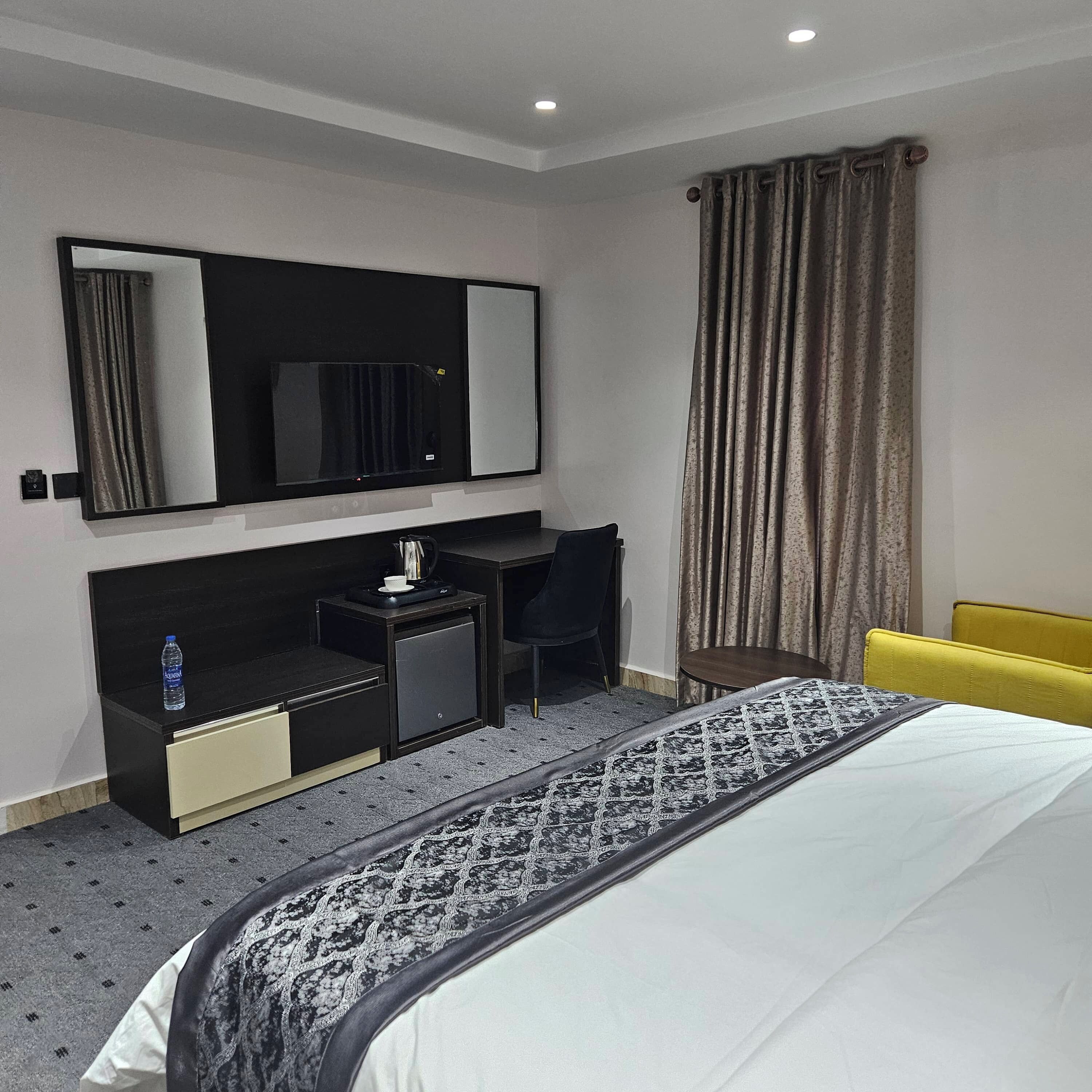 A hotel room with a bed, a desk with a chair, a small cabinet, a mirror, and a window with curtains.