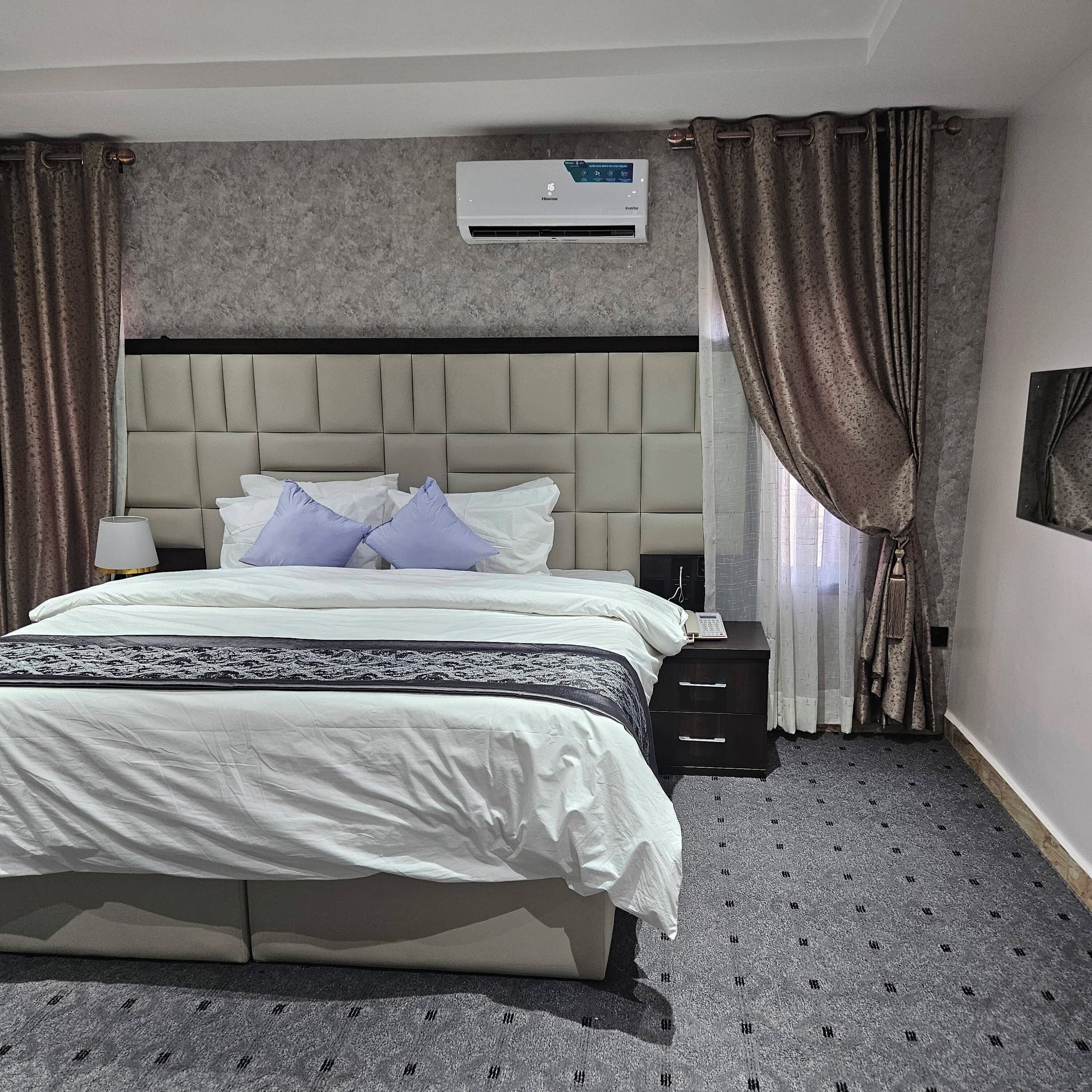 A hotel room with a large bed, a headboard, a nightstand, a lamp, an air conditioning unit, and a window with curtains.