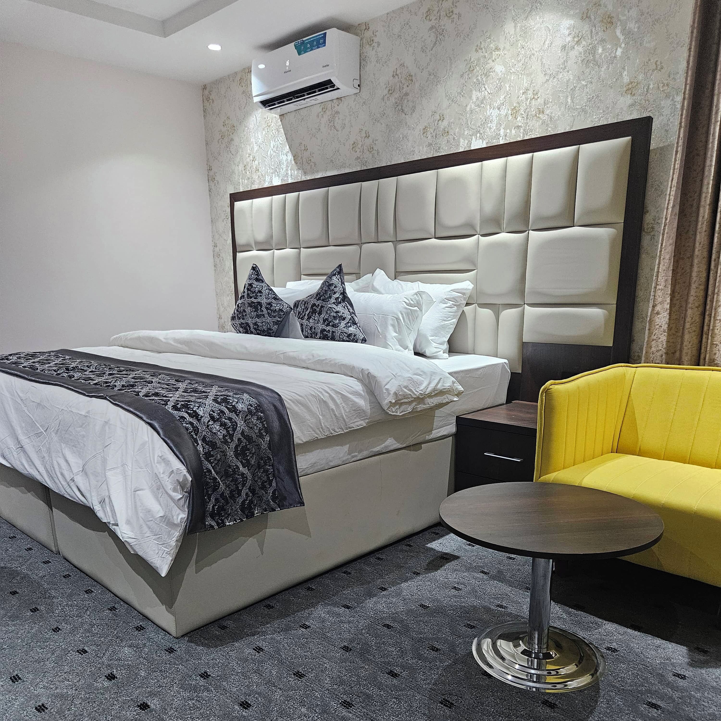 A hotel room with a large bed, a bedside table, a yellow armchair, and an air conditioning unit on the wall.