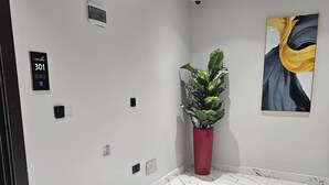 A hallway with a marble floor, a potted plant, a security camera, and a painting on the wall. - Anibex Luxe (Abuja)