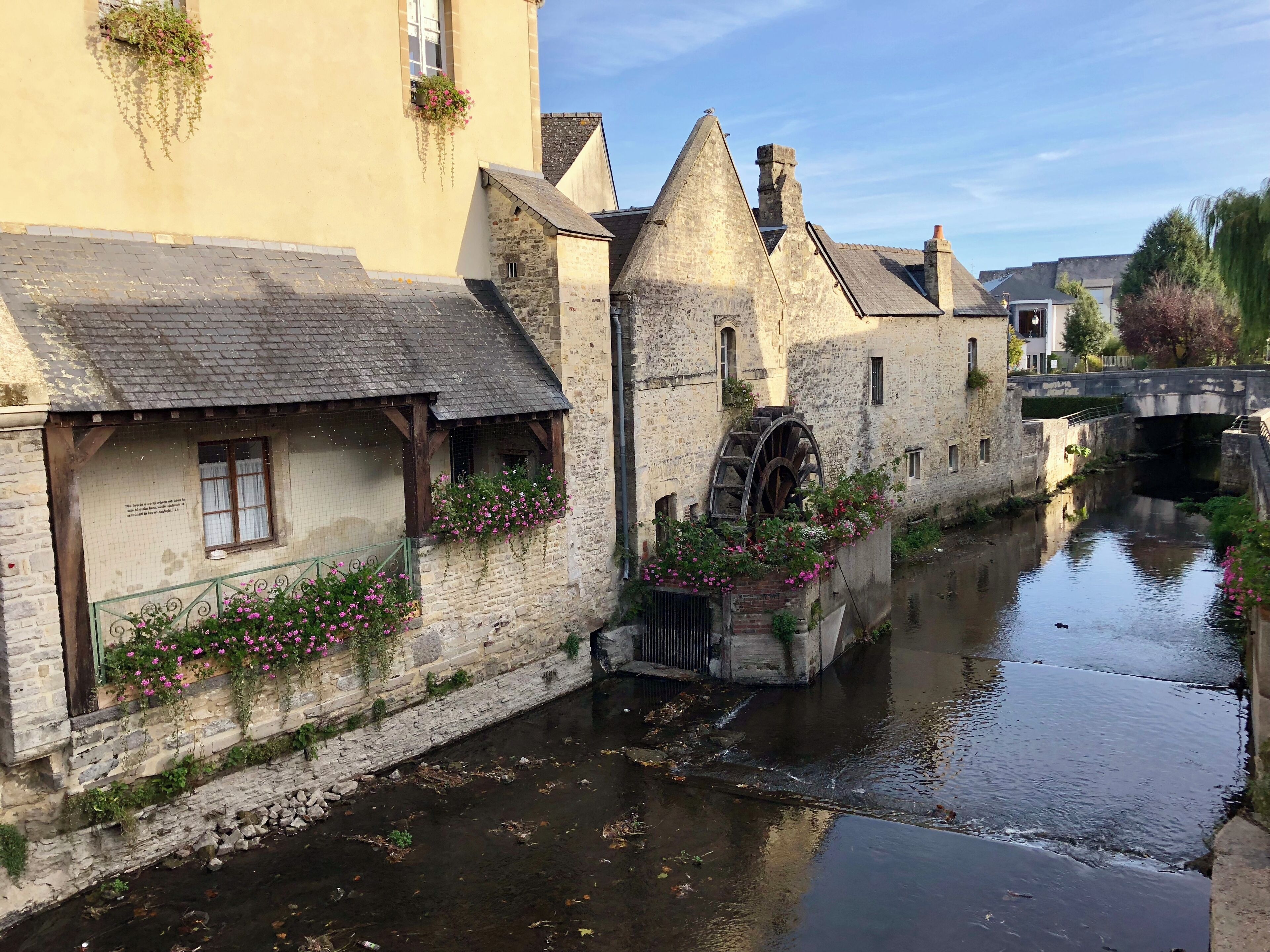 A narrow canal with stone buildings on one side, a water wheel, and a bridge in the background.