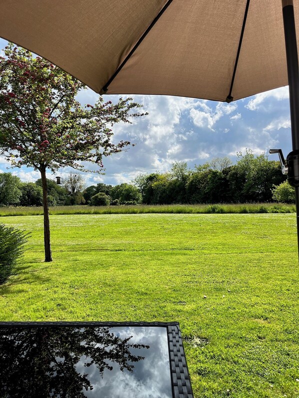 Un patio al aire libre con mesa y sillas bajo un paraguas grande, con vistas a un césped y árboles.