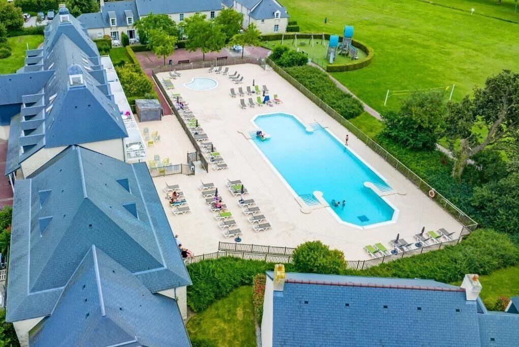En vy från ovan av ett hotell med en stor rektangulär pool, omgiven av solstolar och en gräsyta.