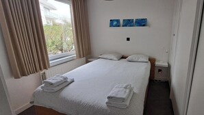 A neatly made bed with two pillows and folded towels, a window with curtains, and three wall-mounted paintings.