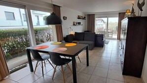 A modern living space with a dining area featuring a table with colorful placemats, a sofa, and a large window overlooking a garden.