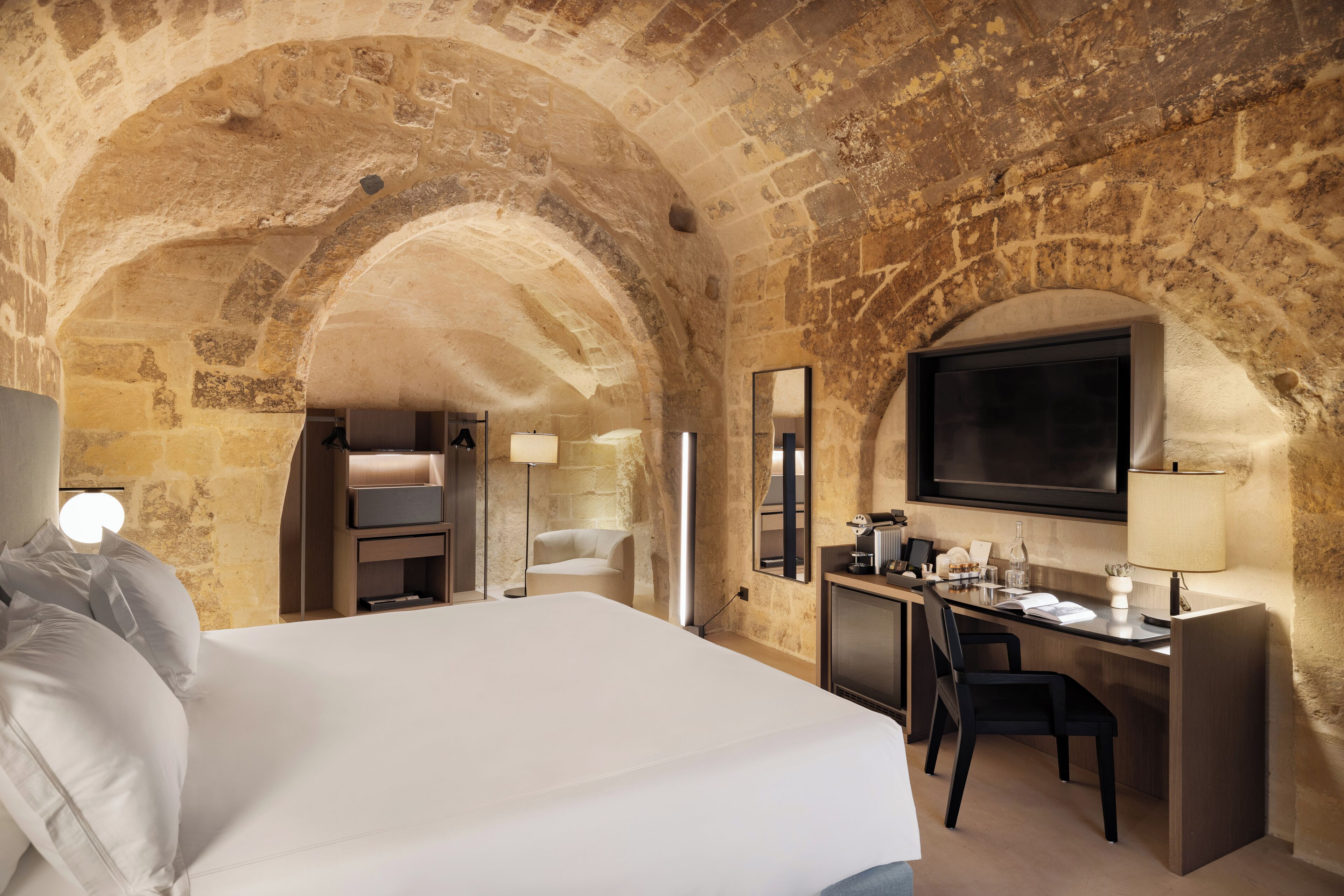 A hotel room with a large bed, a desk, a TV, and stone walls.