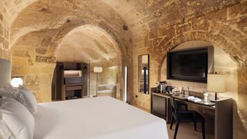 A hotel room with a large bed, a desk, a TV, and stone walls.