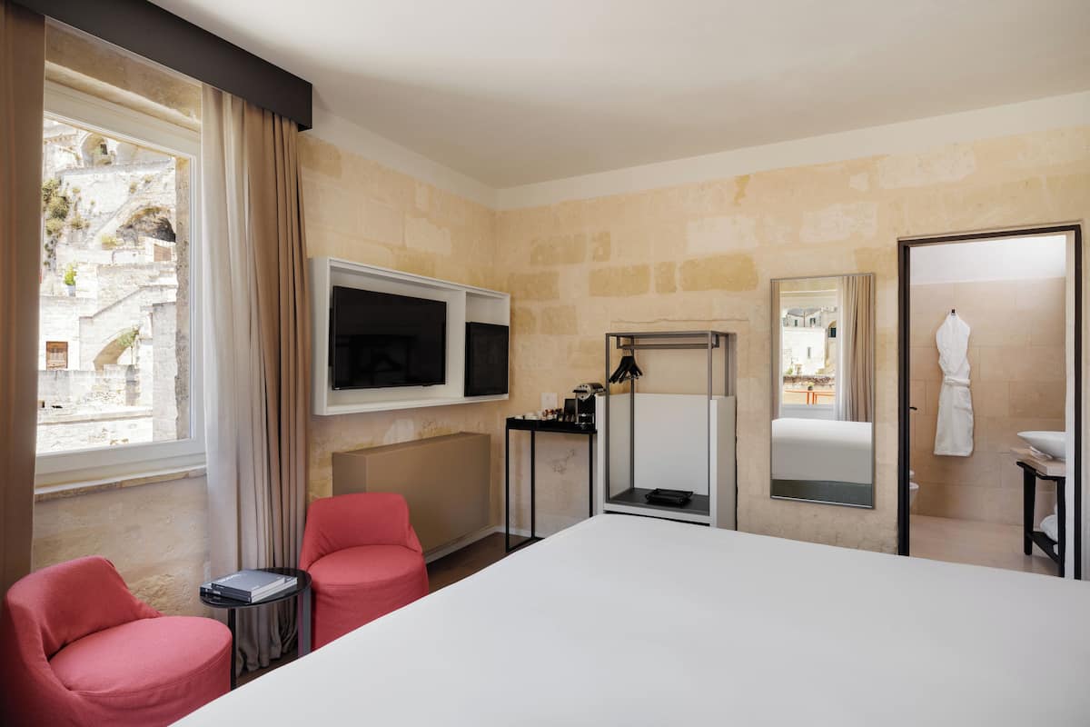 Une chambre d’hôtel avec un grand lit, un téléviseur fixé au mur, une petite table et une chaise. Il y a une fenêtre avec des rideaux, un miroir et un peignoir accroché à la porte.