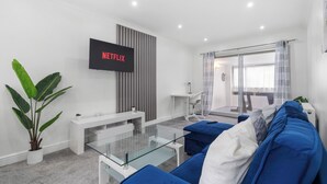 A modern living room with a glass coffee table, blue sofas, a white bench, and a TV mounted on the wall. - Staines House-3bed-sleeps7- Freep-close to Airport (Staines)