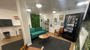 A modern lounge area with a green sofa, wooden chairs, a small table, and a vending machine. - Student Only Apollo Court (Liverpool)