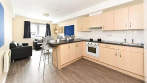 A modern kitchen with light wood cabinets, a black countertop, and a built-in oven. There is a dining area with a table and chairs. - Student Only Apollo Court (Liverpool)