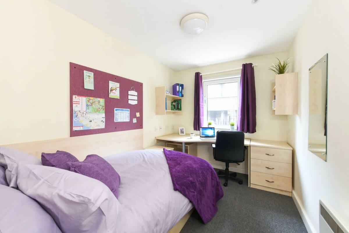 Une chambre d’hôtel avec un lit, un bureau avec un ordinateur, une fenêtre avec des rideaux et un tableau d’affichage au mur.