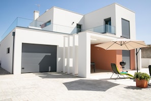Una casa moderna de dos pisos con garaje, un balcón con barandilla de vidrio y un patio con una silla y sombrilla.