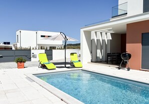 Une piscine avec deux chaises longues, un barbecue et un bâtiment moderne doté d’un toit plat.