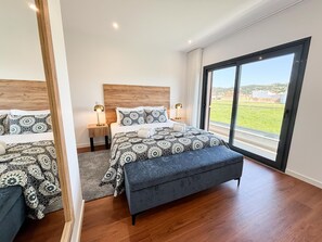Un dormitorio con un ventanal grande, un cabecero de madera, una cama con ropa de cama estampada, un banco y una mesita de noche con lámparas.