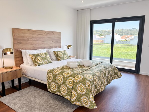 A bedroom with a large bed, a nightstand with a lamp, a view of the outdoors through a window, and a wooden headboard.