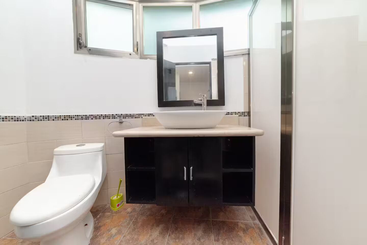 A bathroom with a white toilet, a black cabinet with a mirror above the sink, and a window above the sink.