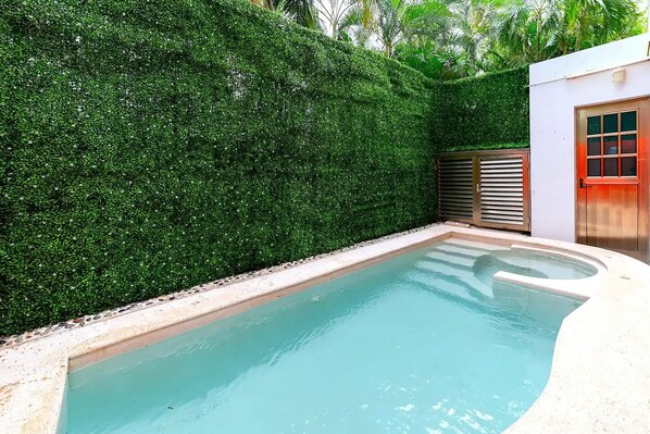 A swimming pool with a surrounding hedge, a door with a red frame, and a small building with a window.