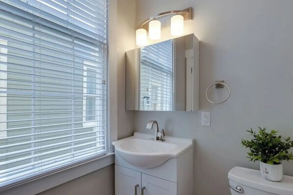 Ein Badezimmer mit einem weißen Waschbecken, einem Spiegel und einer Toilette. Ein Fenster mit Jalousien und ein Blumentopf stehen auf der Toilette.