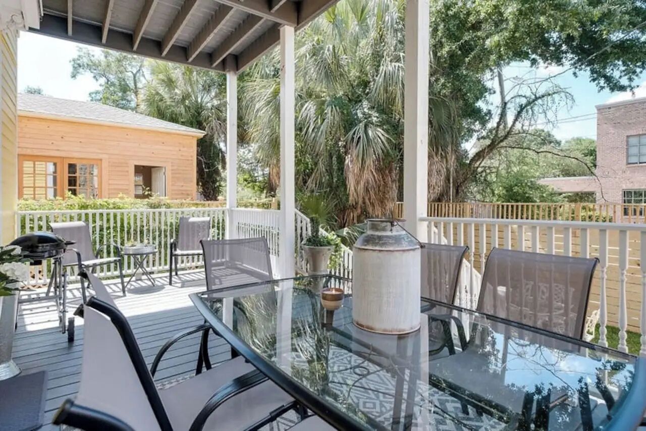 A covered outdoor patio with a glass table and chairs, surrounded by a white fence and palm trees.