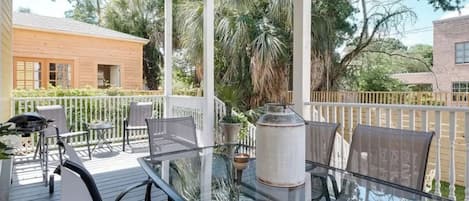 A covered outdoor patio with a glass table and chairs, surrounded by a white fence and palm trees.