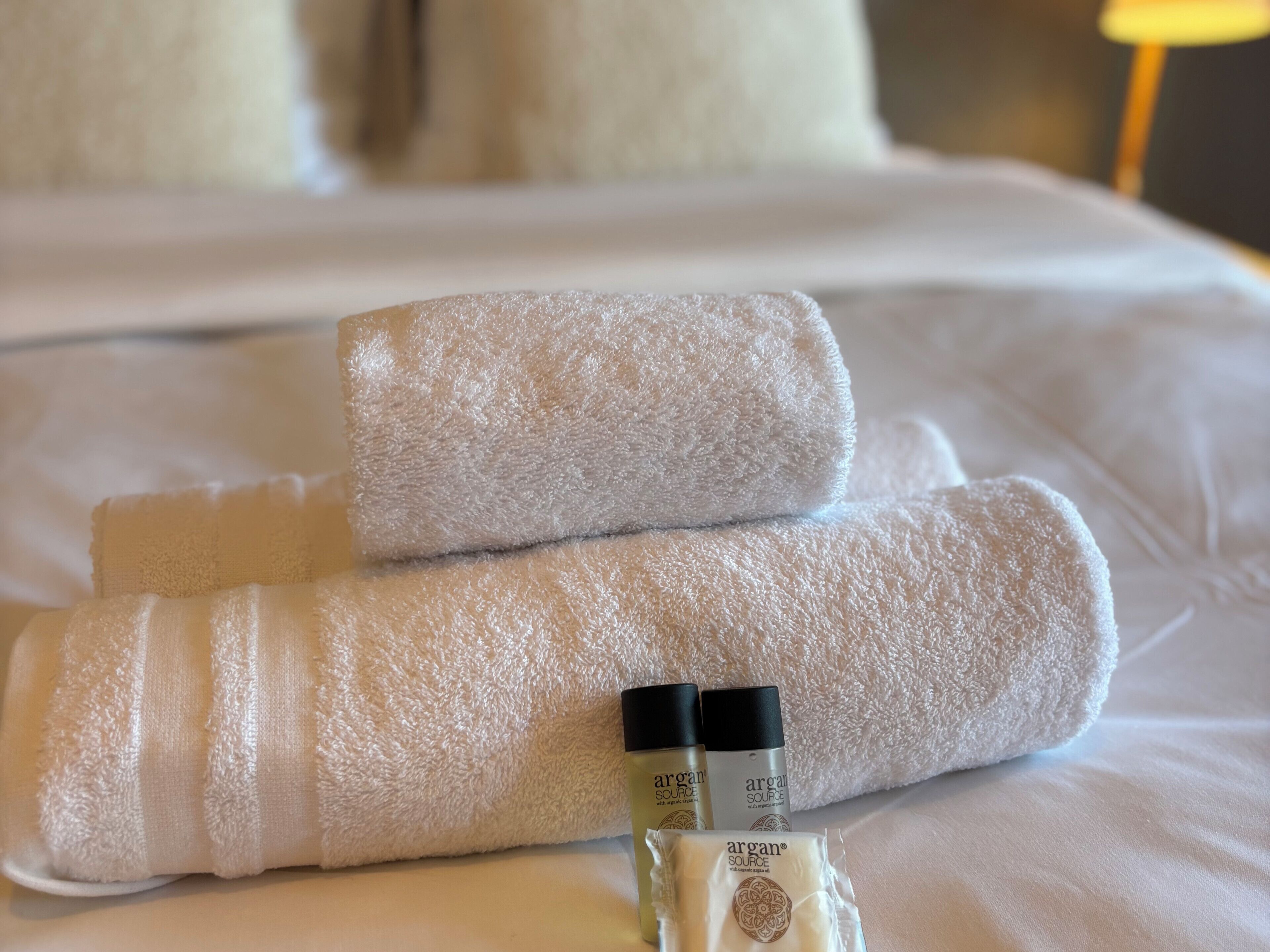 Ein ordentlich gemachtes Bett mit zwei aufgerollten Handtüchern und zwei Flaschen Argan Source Lotion.