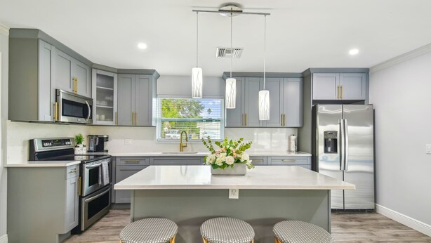 A modern kitchen with grey cabinets, stainless steel appliances, a central island with stools, pendant lighting, and a window providing a view of the outdoors. - Tropical Oasis •Pet-Friendly + Pool Clearwater (Largo)