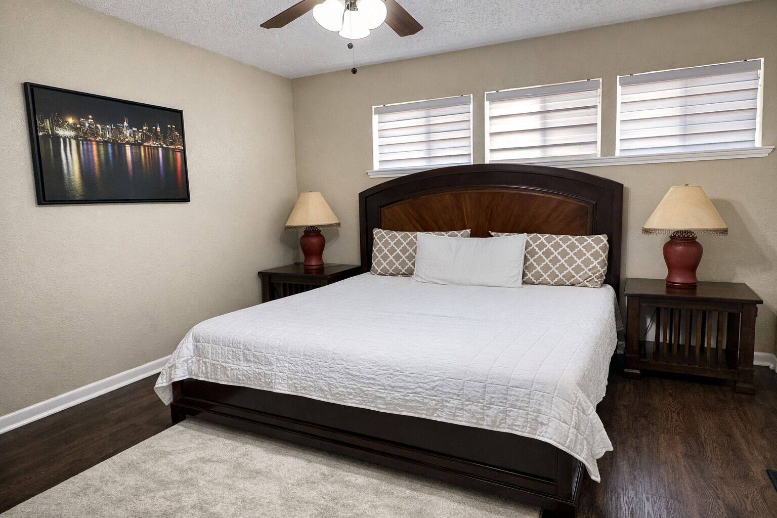 A bedroom with a large bed, two bedside tables with lamps, a ceiling fan, and a cityscape painting on the wall.