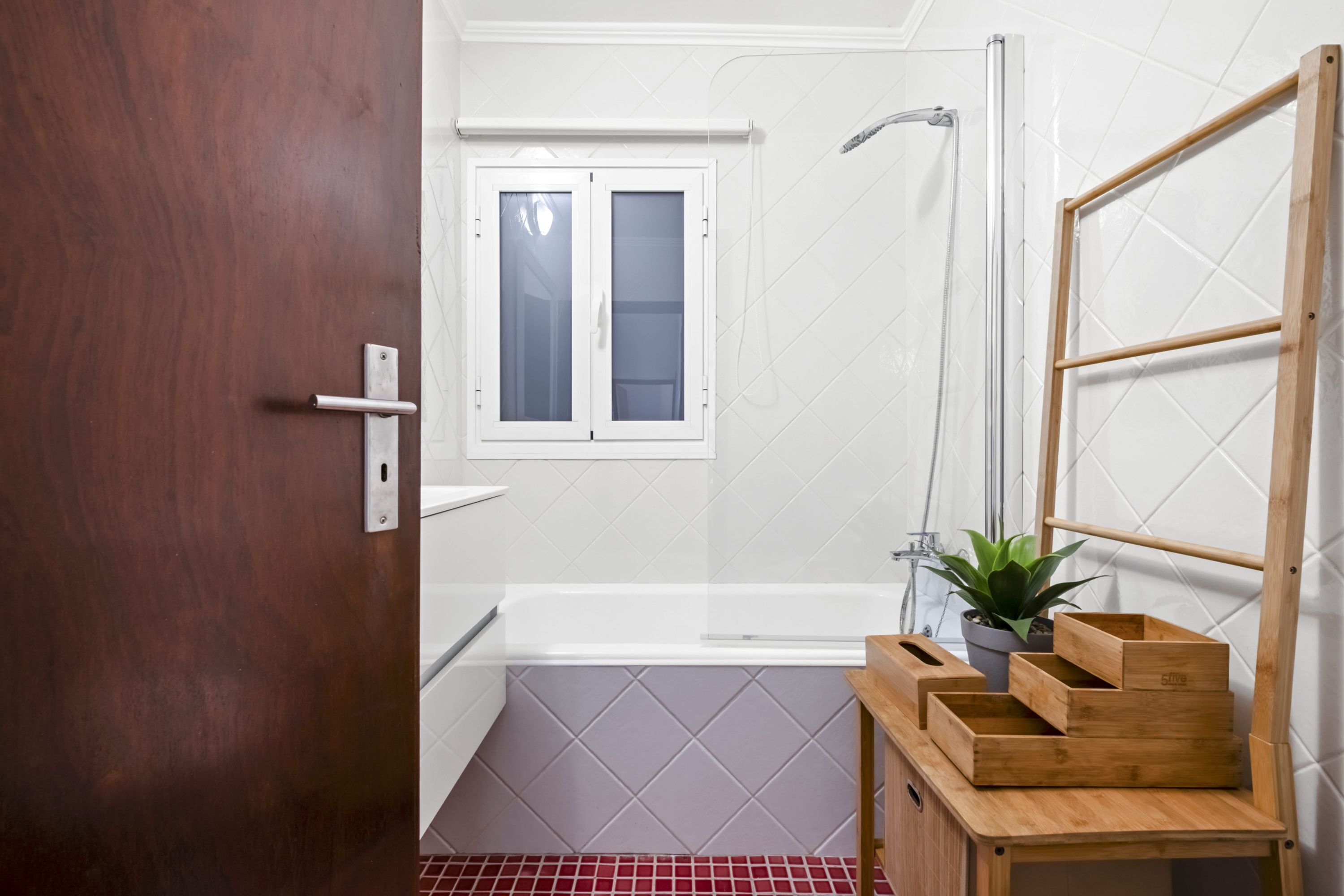 A bathroom with a wooden door, a white bathtub, a shower, a window, and a wooden shelf with a potted plant.