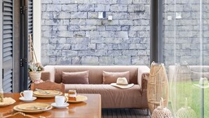 Un coin repas chaleureux avec une table en bois dressée pour deux personnes, un canapé beige agrémenté d'un chapeau, des lampes en osier et, en arrière-plan, un mur en pierre.