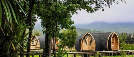 A rustic cabin with a wooden deck, surrounded by lush greenery and a mountainous backdrop.